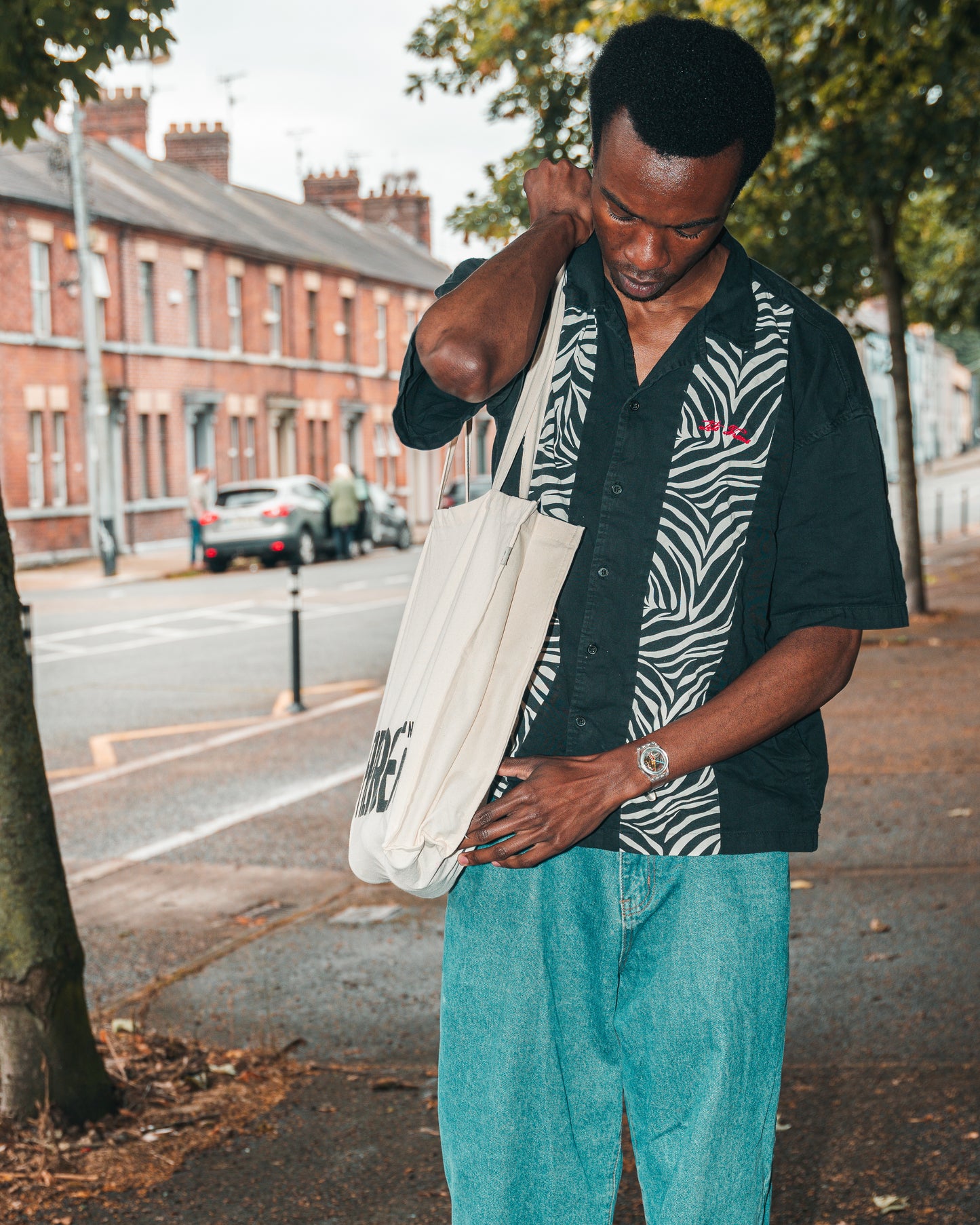Model is shown walking on pavement close to a terrace of red brick houses wearing the ecru Fierce Mild non-alc tote bag on right shoulder, with blue jeans, a black and white shirt with a zebra pattern on the lapels, and a wrist watch.