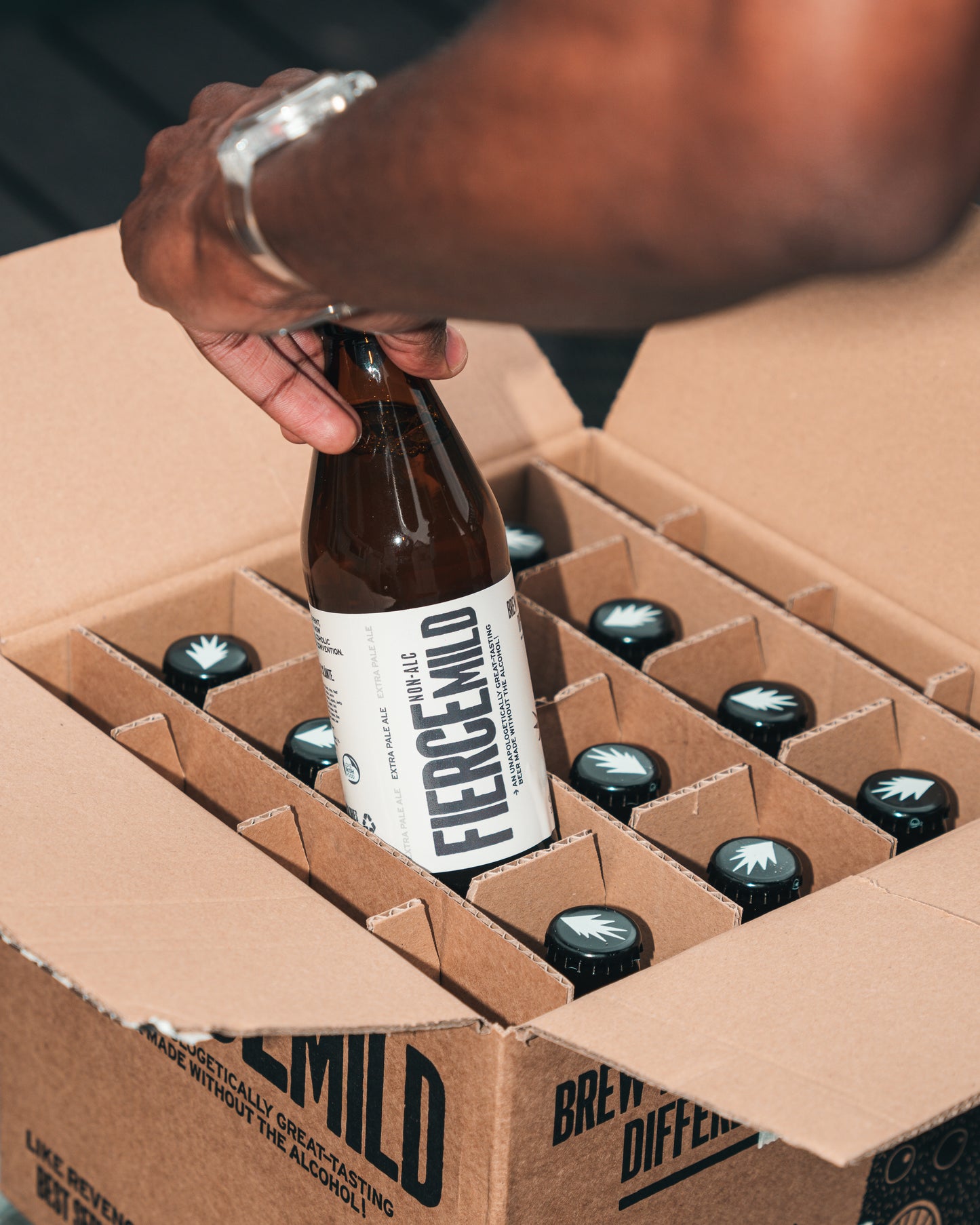 Arm with wrist watch pulls a brown glass bottle of Fierce Mild non-alc beer from a craft paper brown box that houses 15 other brown glass bottles of Fierce Mild non-alc beer.