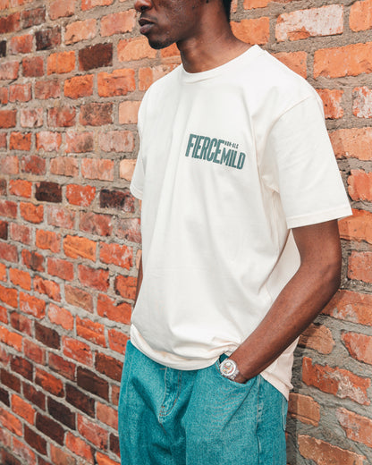 Model showcases ecru Fierce Mild non-alc beer merchandise t-shirt. Model is shown from the side against a red brick wall, and pairs the t-shirt with loose fitting blue jeans, and a wrist watch with a clear strap.