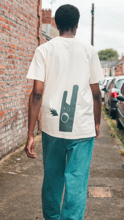 Model showcases ecru Fierce Mild non-alc beer merchandise t-shirt. Model is shown from behind and walking on the pavement. The t-shirt shows the Fierce Mild character in black. The model pairs the t-shirt with loose fitting blue jeans, and a wrist watch with a clear strap.