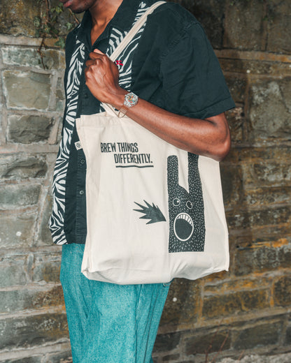 Model is shown to the side and alongside a stone wall wearing the ecru Fierce Mild non-alc tote bag on left shoulder, with blue jeans, a black and white shirt, and a wrist watch with a clear strap. The tote bag shows the tagline Brew Things Differently and the character breathing fire in black.