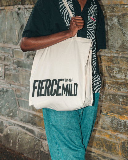 Model is shown wearing the ecru Fierce Mild non-alc tote bag on right shoulder, with blue jeans, and a black and white shirt, and walks alongside a stone wall. 