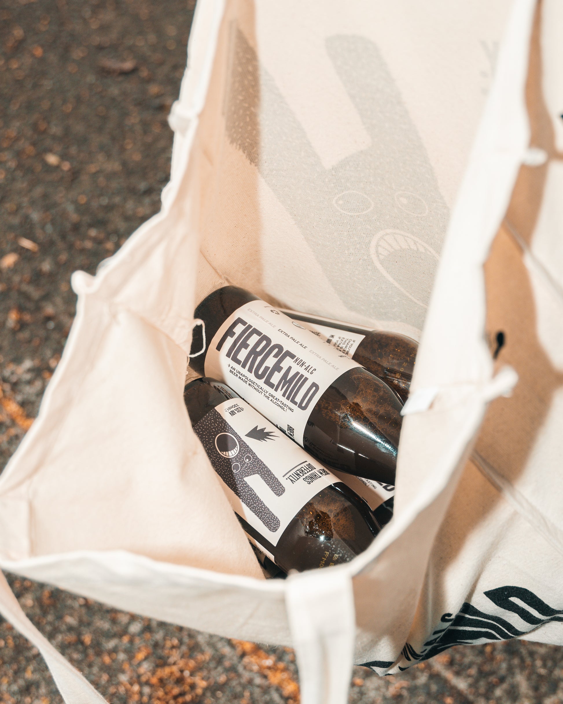 Ecru Fierce Mild non-alc tote bag is open on one side, and shows brown glass branded bottles of Fierce Mild non-alc beer inside. 