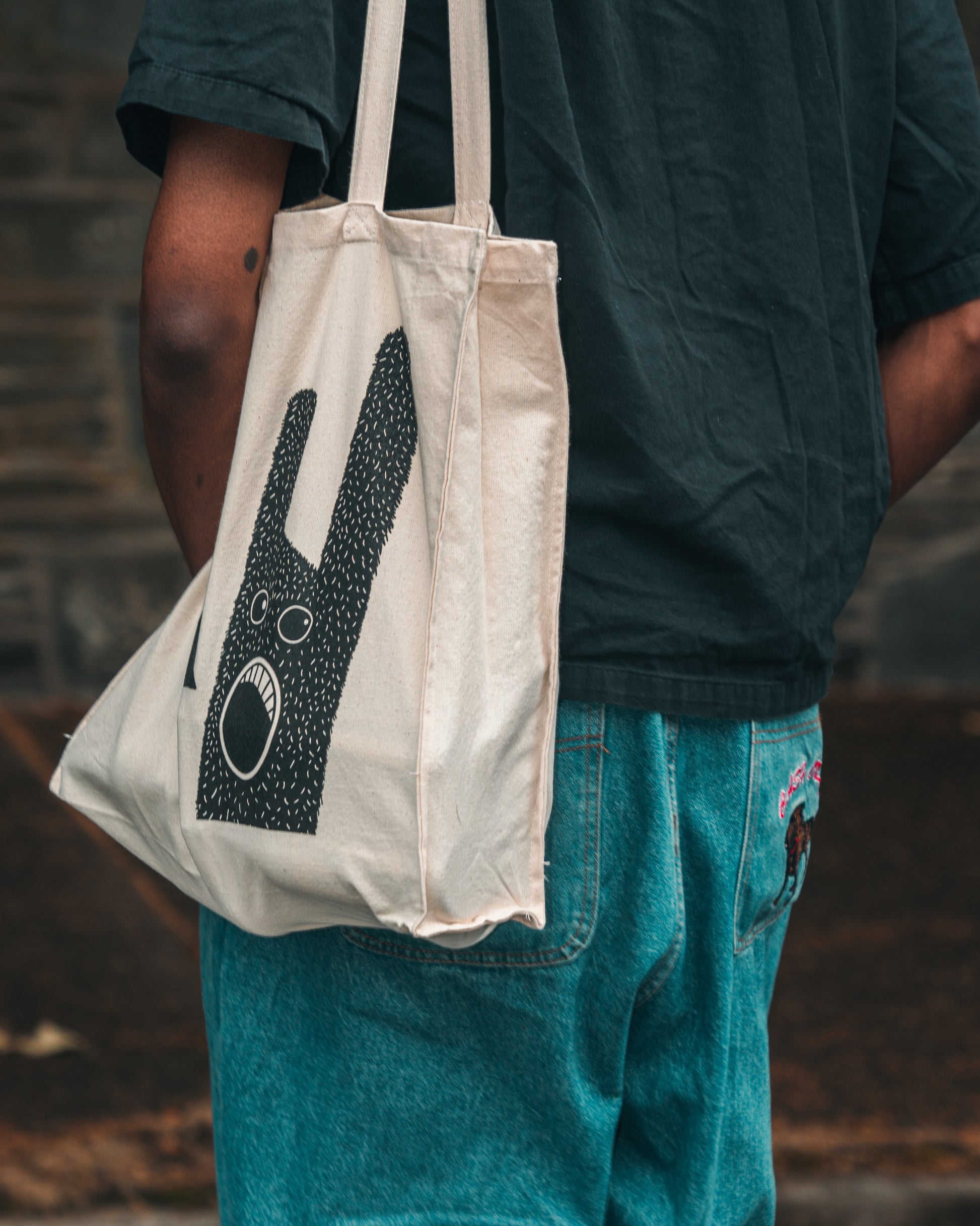 Model is shown from behind wearing the ecru Fierce Mild non-alc tote bag on left shoulder, with blue jeans, and a black shirt. The tote bag shows the character in black.