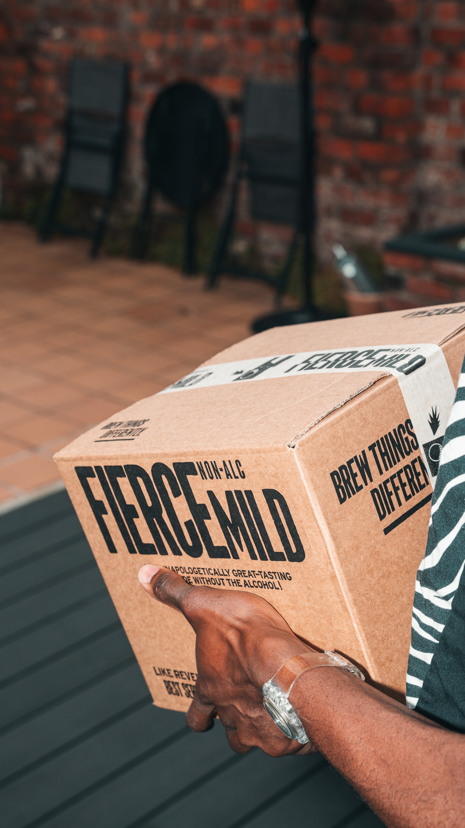 Arm with wrist watch is shown holding Fierce Mild non-alc beer packaged in a branded craft brown box with white branded label tape.